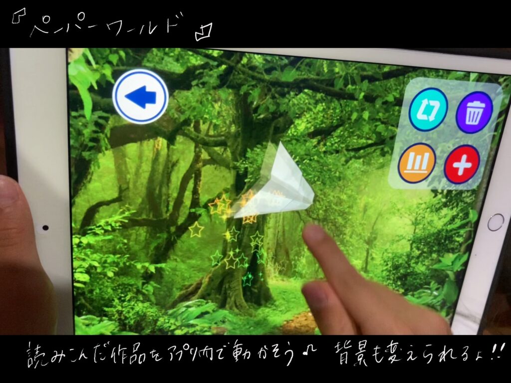 ペーパーワールドのアプリで遊んでいる写真。左手でアイパットを支えて、右手人差し指で、画面を操作している。画面には森の風景。その中に自分でつくった折り紙の紙飛行がある。
