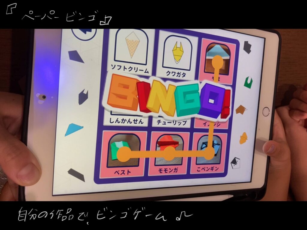 ペーパーラボの体験アプリでビンゴゲームをしている写真。子供がアイパットを両手で持っている。画面人は折り紙で作った、ベスト、モモンガ、こペンギンで横のビンゴが完成している。山、イノシシ、こペンギンで縦のビンゴが完成している。画面中央には、カラフルな文字でBINGOと書いてある。