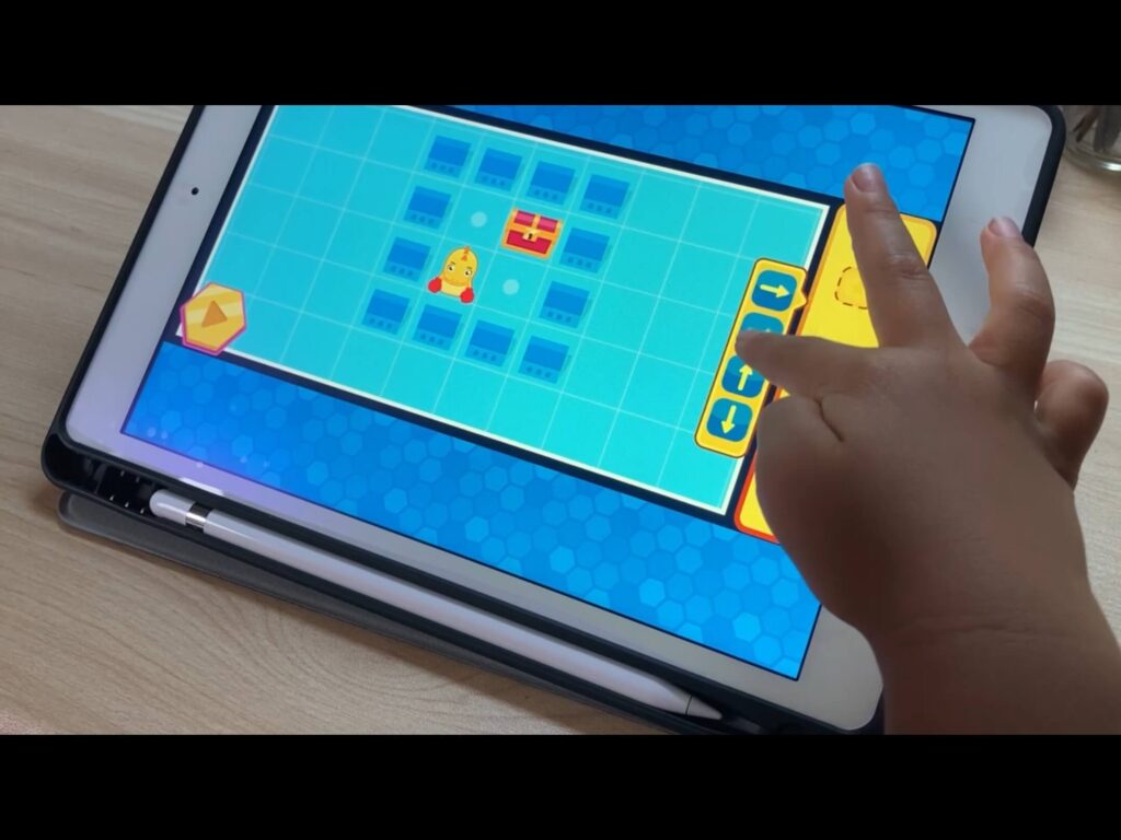 体験版小学生向けアプリ、コードクラフターズで遊んでいる様子。青い背景に、恐竜のキャラクーと宝箱。右には4方向の矢印のイラスト。子供の手が矢印をタップしている。