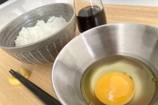 木製のトレイに朝食が並べられている写真。右に、生卵が入った柳宗理のステンレスボール13cm。
左には、グレーの茶碗に盛られた白米と醤油さしが置かれている。ボウルと茶碗の下には、黒い箸と黄色い箸置きが添えられている。