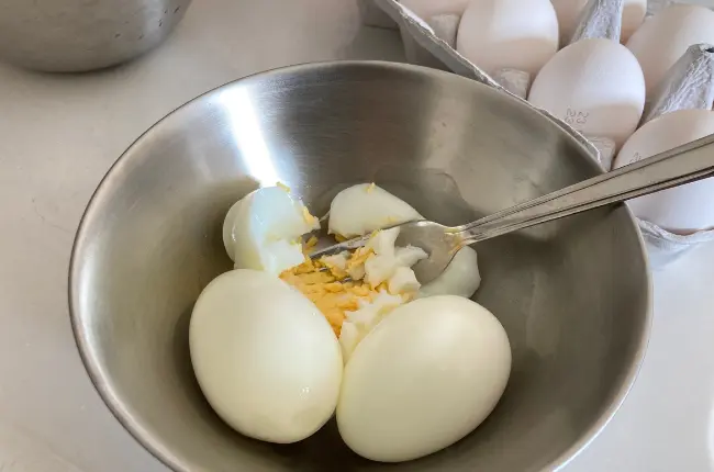 調理中の写真。柳宗理のステンレスボウル16cmに、殻をむいたゆでたまご3つ入っている。3つの内1つの卵は、フォークで潰されている。ボウルの後ろには、生卵のパックが置かれている。