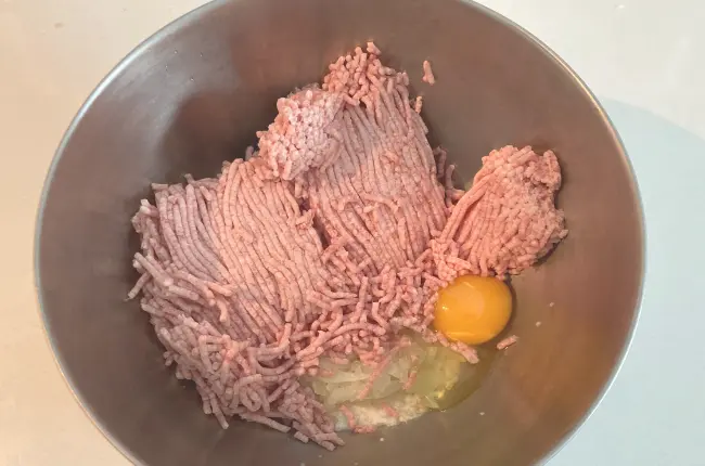 白いキッチントップの背景。ハンバーグの具材を上から撮った写真。柳宗理のステンレスボール23cmの中にひき肉約600gと生卵とパン粉が入っている。